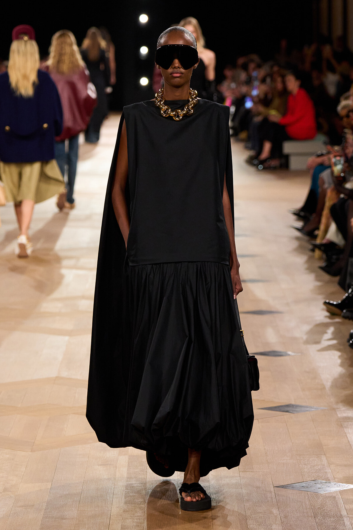 Model on runway wearing black sleeveless cape top and voluminous skirt, oversized sunglasses, chunky gold necklace, and sandals.