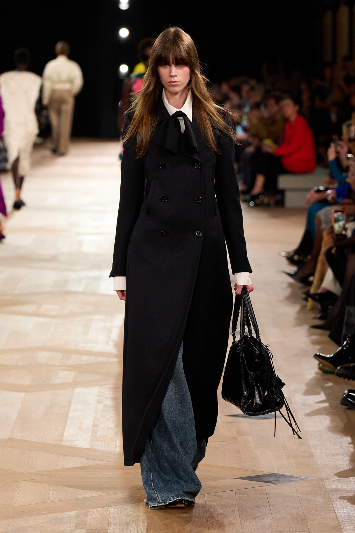 Model walking a runway in a long black double-breasted coat over white-citted shirt, paired with wide-leg blue jeans and a black handbag with tassels.