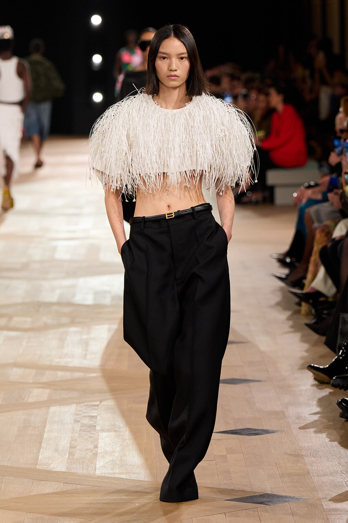 Model walks a runway in a white fringe crop top and black high-waisted wide-leg pants with a belt.