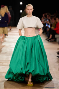 Model on a runway wearing a cream cropped top and a voluminous emerald-green ball gown, walking toward the camera.