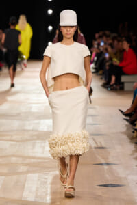 Model walking a runway in a white cropped top, high-waisted skirt with a floral-midi hem and matching cap, beige sandals.