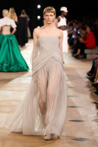 Model walks the runway in a pale gray, strapless, draped gown with a sheer overlay and flowing skirt.
