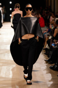 Model strides the runway in a black cape top, black trousers, oversized sunglasses, and chunky gold jewelry.
