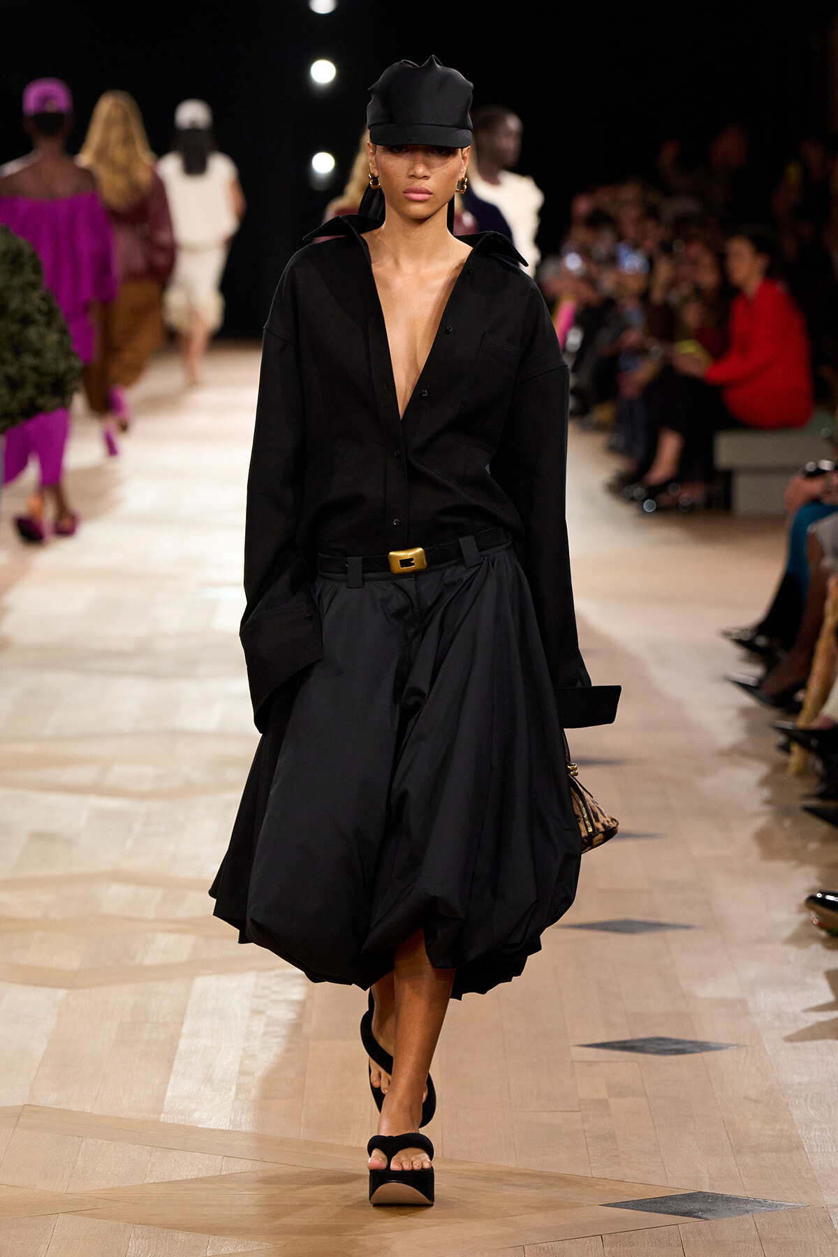 Runway model in a black oversized blouse and dramatic billowing black skirt, wearing a black cap and platform sandals.