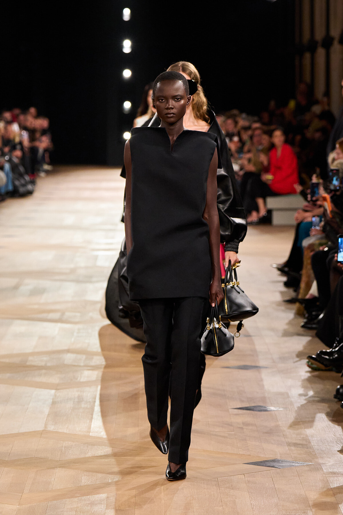 Model on a runway wearing an all-black outfit: sleeveless, boxy tunic and tailored pants, carrying a black handbag with gold hardware.
