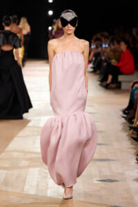 Model walks the runway in a strapless pale pink gown with a voluminous bubble skirt and a jeweled black visor-style headpiece.