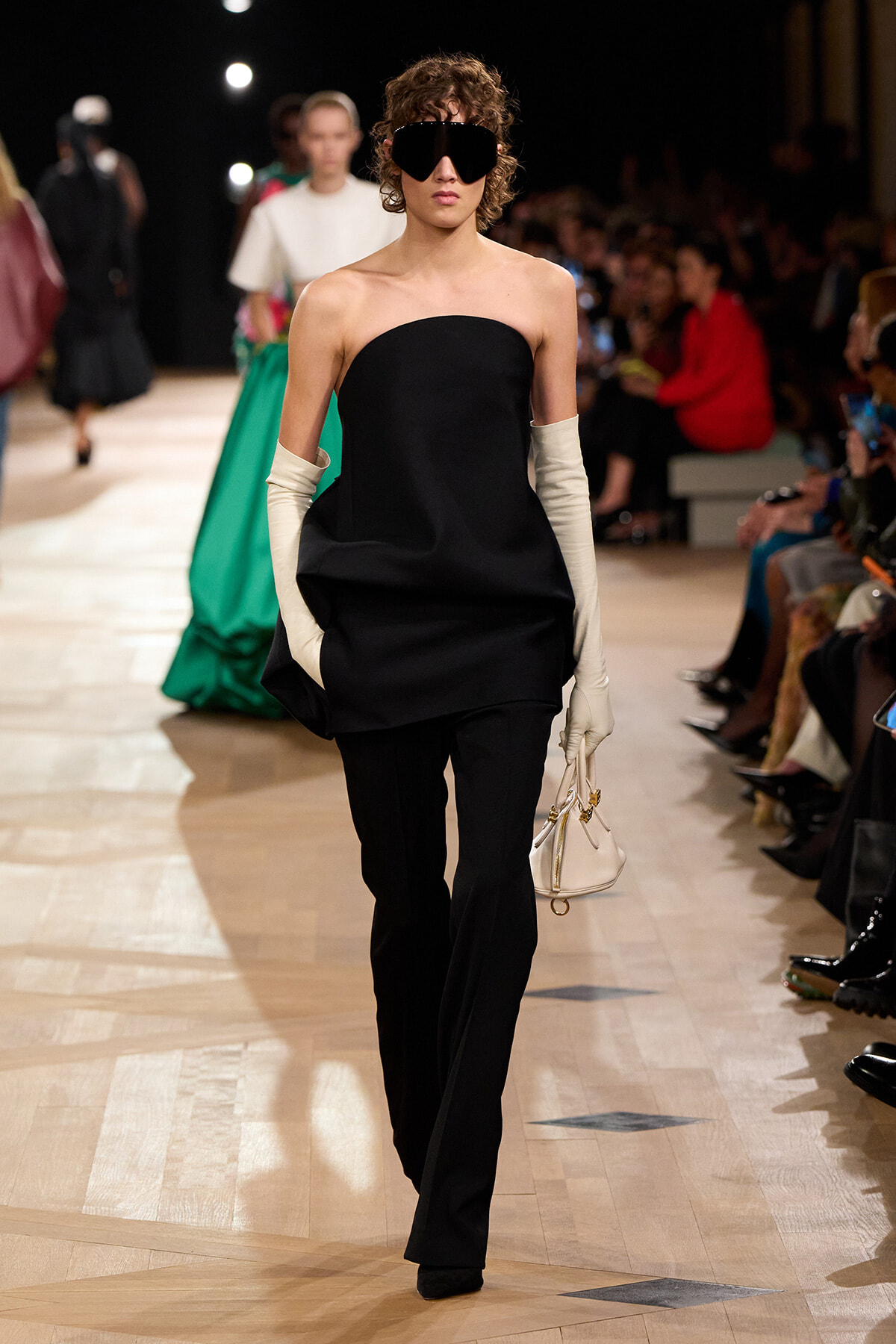 Runway model in a strapless black top and black pants, wearing oversized sunglasses and cream opera gloves, carrying a small white handbag.