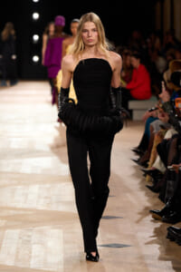 Model walks a fashion runway in a black strapless jumpsuit with oversized ruffled sleeves and long black gloves.