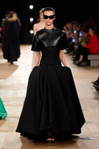 Model walks a fashion runway in a matte black leather top with a full, voluminous black skirt; futuristic sunglasses and minimal footwear visible on a wooden floor runway.