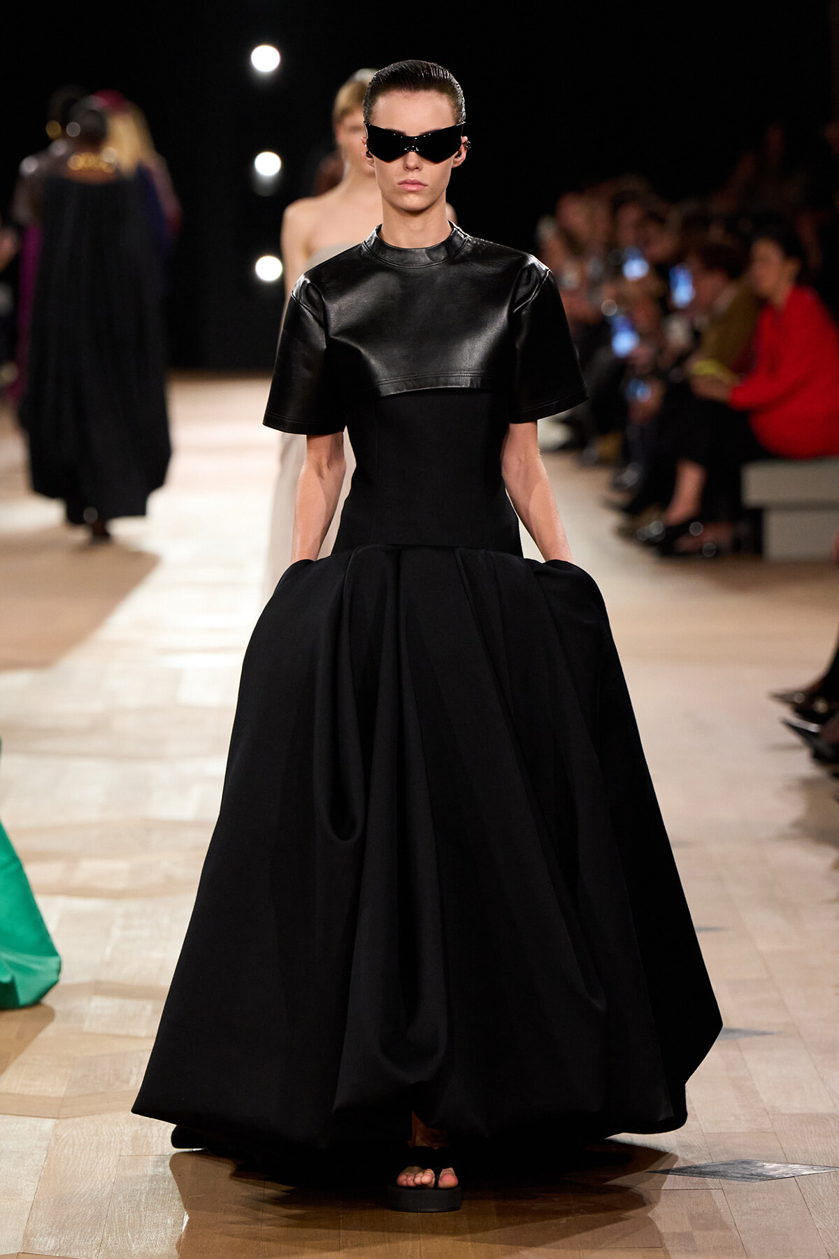 Model walks a fashion runway in a matte black leather top with a full, voluminous black skirt; futuristic sunglasses and minimal footwear visible on a wooden floor runway.