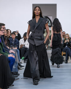 Model in a dark, patchwork-style suit walks the runway as an audience watches.