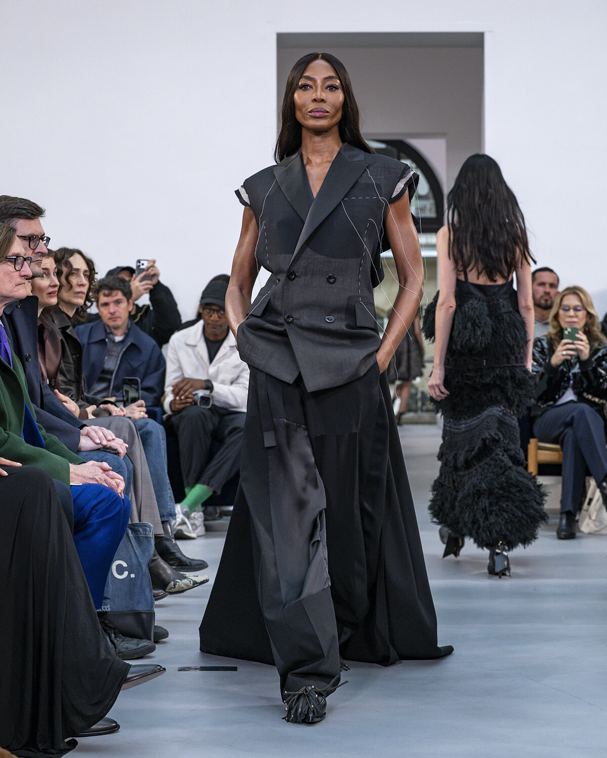 Model in a dark, patchwork-style suit walks the runway as an audience watches.