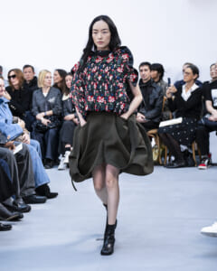 Model walks the runway in a dark floral blouse with puffed sleeves and a large olive bubble skirt, black ankle boots, as an audience watches.