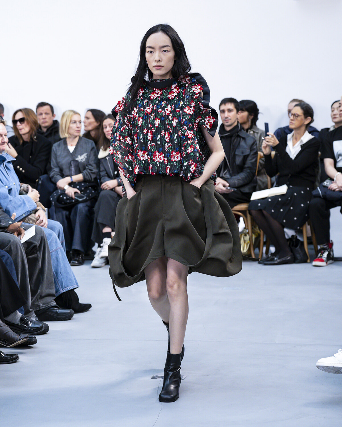 Model walks the runway in a dark floral blouse with puffed sleeves and a large olive bubble skirt, black ankle boots, as an audience watches.