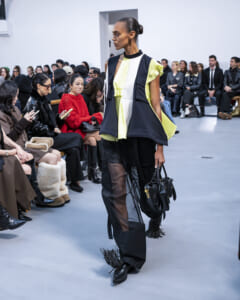 Model walks the runway in a bold color-blocked lime, white, and navy outfit with sheer-panel pants, holding a black bag as the audience watches.