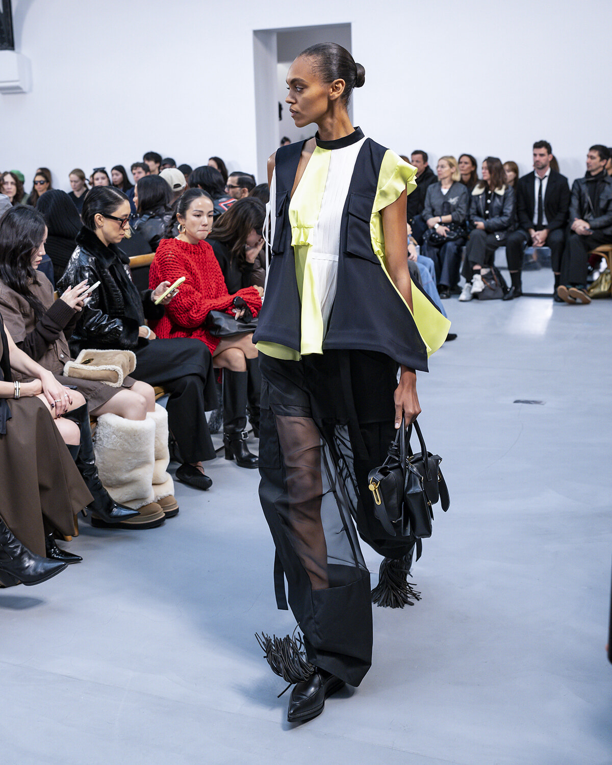 Model walks the runway in a bold color-blocked lime, white, and navy outfit with sheer-panel pants, holding a black bag as the audience watches.