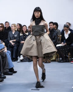Runway model walking in a fashion show wearing a voluminous beige strapless dress with a black neck scarf and chunky black fringed shoes, audience in background.