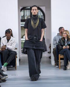 Model walks a fashion runway in a black top with a bold gold fringe necklace and layered dark denim; audience seated along the sides.