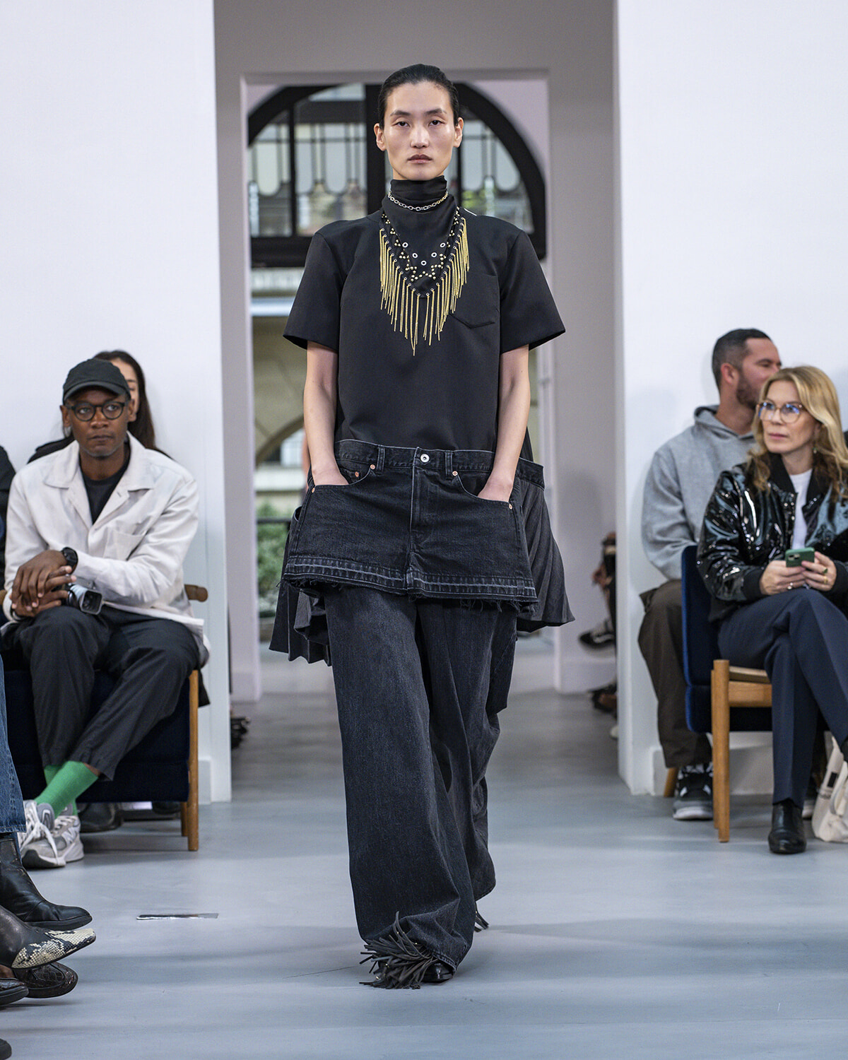 Model walks a fashion runway in a black top with a bold gold fringe necklace and layered dark denim; audience seated along the sides.