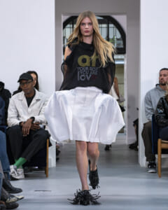 Model walks a runway in a black mesh-shoulder top and a large white sculpted skirt, surrounded by seated audience.
