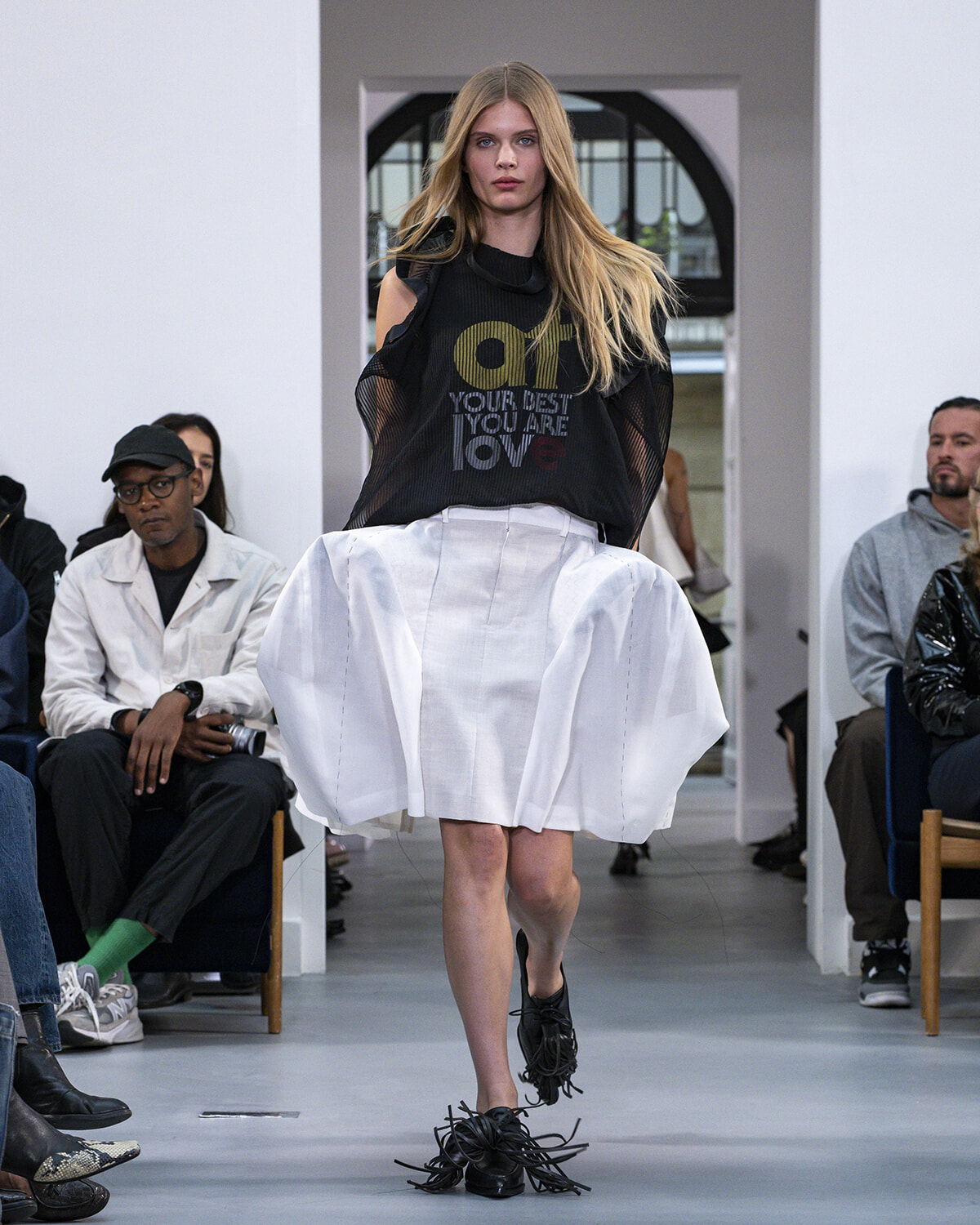 Model walks a runway in a black mesh-shoulder top and a large white sculpted skirt, surrounded by seated audience.