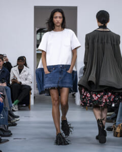 Model walks runway in a white oversized top over a denim mini skirt with fringed black shoes; audience watches from benches on sides.