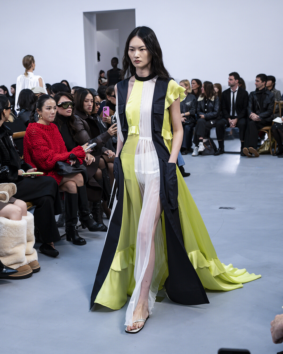 Model walks the runway in a color-block yellow and navy coat with sheer white panels, audience watching from the sides.