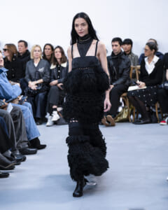 Model walking a runway in an all-black, textured fringe dress with sheer panels and ankle boots, audience watching.