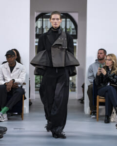 Model walks a fashion runway in a layered black and gray avant-garde outfit with oversized sleeves and fringe at the pants. Audience sits along the aisle, watching.