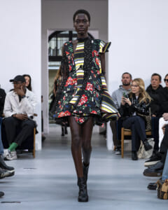 Model walks a runway in a bold, mixed floral and striped dress with black ankle boots; audience seated along the sides.
