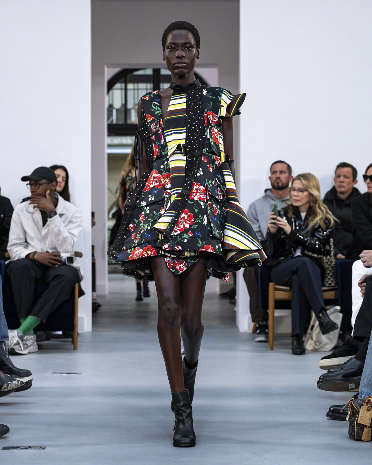 Model walks a runway in a bold, mixed floral and striped dress with black ankle boots; audience seated along the sides.