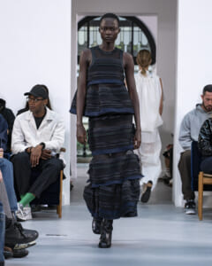 Model walking a dark, tiered striped dress on a runway with audience seated along both sides of the catwalk.
