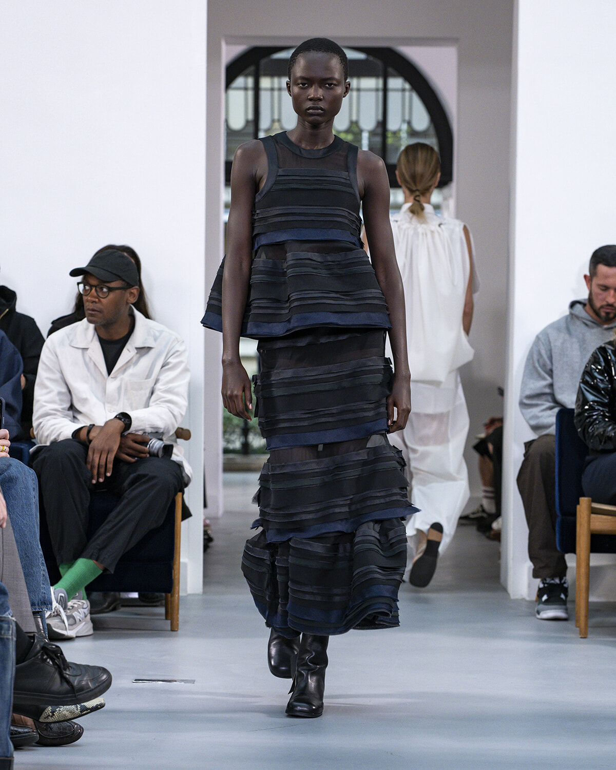 Model walking a dark, tiered striped dress on a runway with audience seated along both sides of the catwalk.