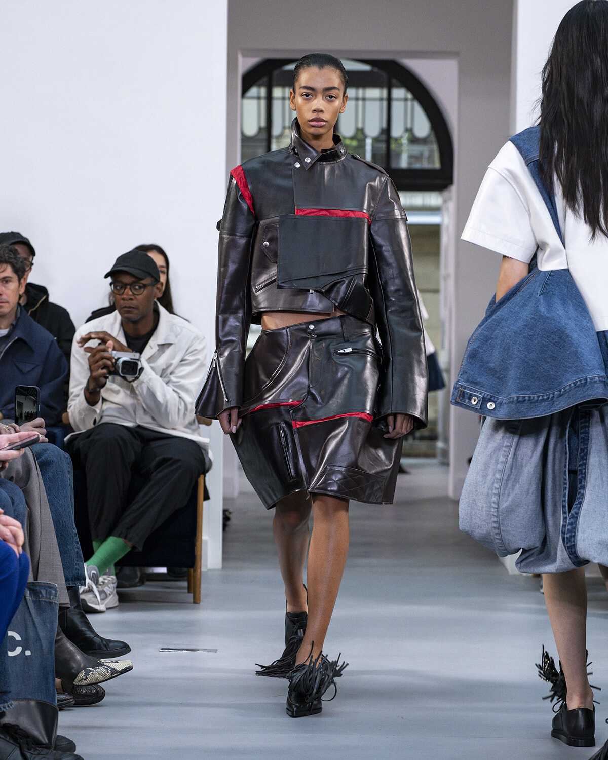 Model walks the runway in a dark leather outfit with red piping—a cropped top and knee-length skirt with pockets; seated audience and a photographer visible in the background.
