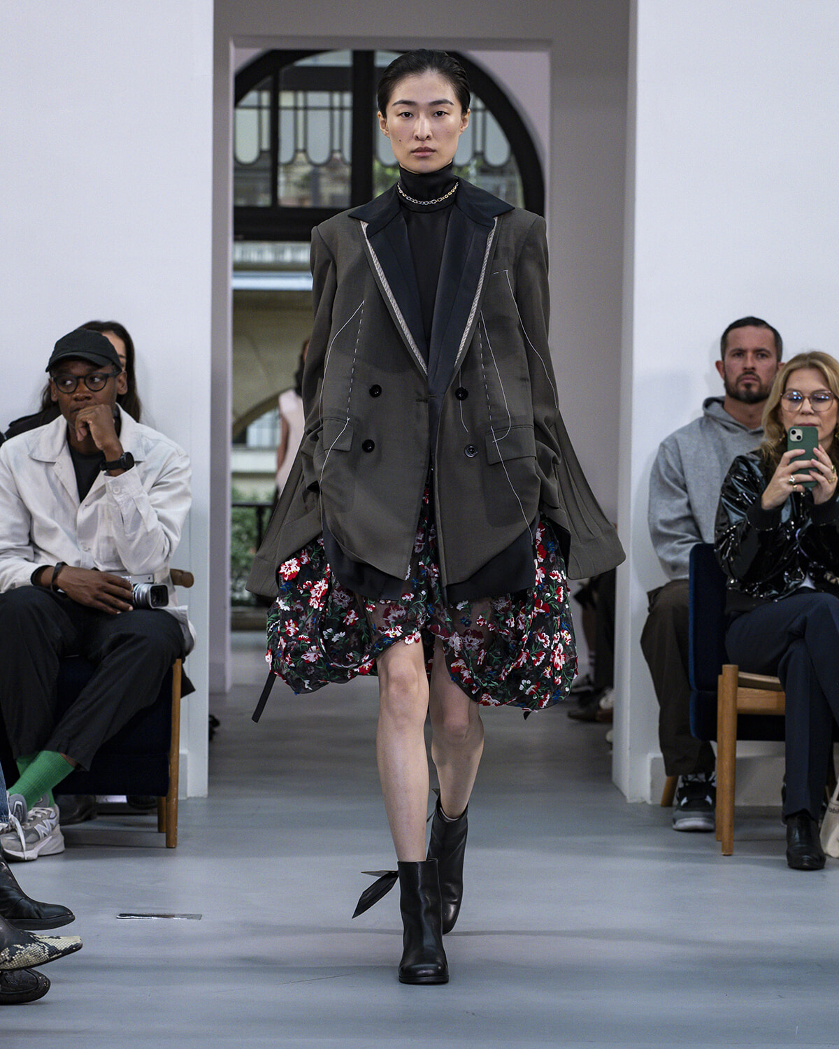 Model walks a fashion runway in an oversized olive blazer with contrast piping, a black turtleneck, and a floral bubble skirt with black boots, audience in background.