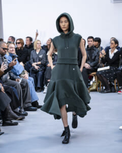 Model wearing a dark green hooded knit dress with a wide ribbed belt and ankle boots walks the runway.