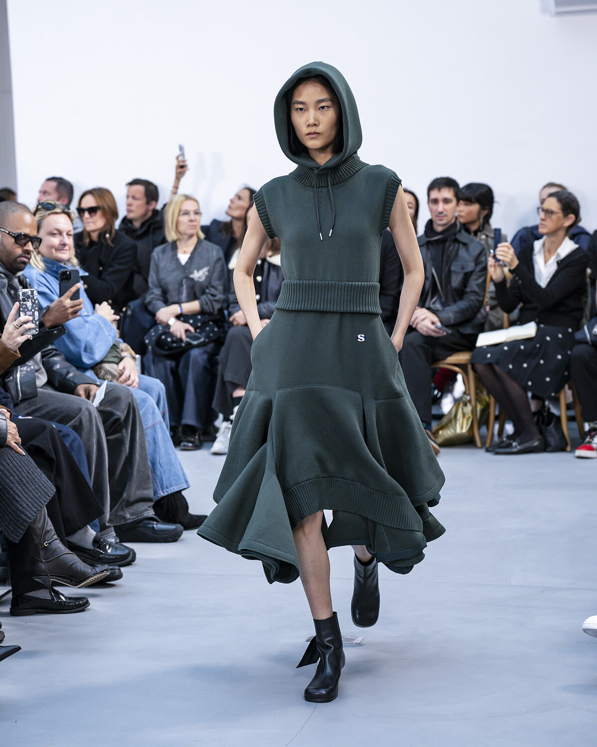 Model wearing a dark green hooded knit dress with a wide ribbed belt and ankle boots walks the runway.