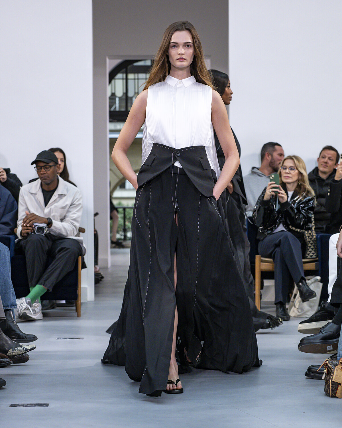 Model walking the runway in a white sleeveless blouse paired with a black high-waisted wide-leg skirt featuring a structured, oversized waist; seated audience watches.