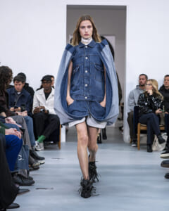 Model walking a fashion runway in a denim layered outfit: oversized denim jacket draped over shoulders, white top, and fringe-detail shoes, audience watching.