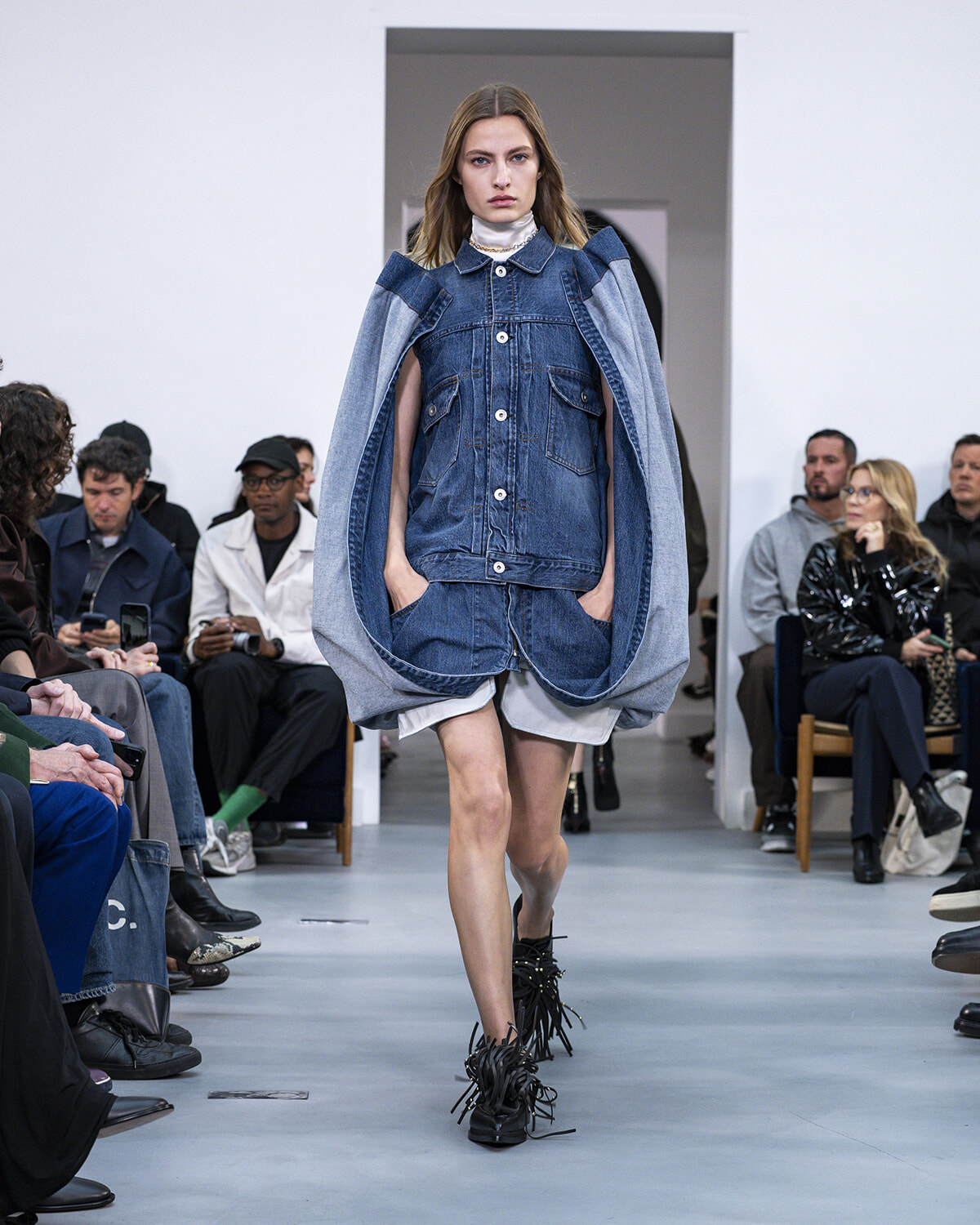 Model walking a fashion runway in a denim layered outfit: oversized denim jacket draped over shoulders, white top, and fringe-detail shoes, audience watching.
