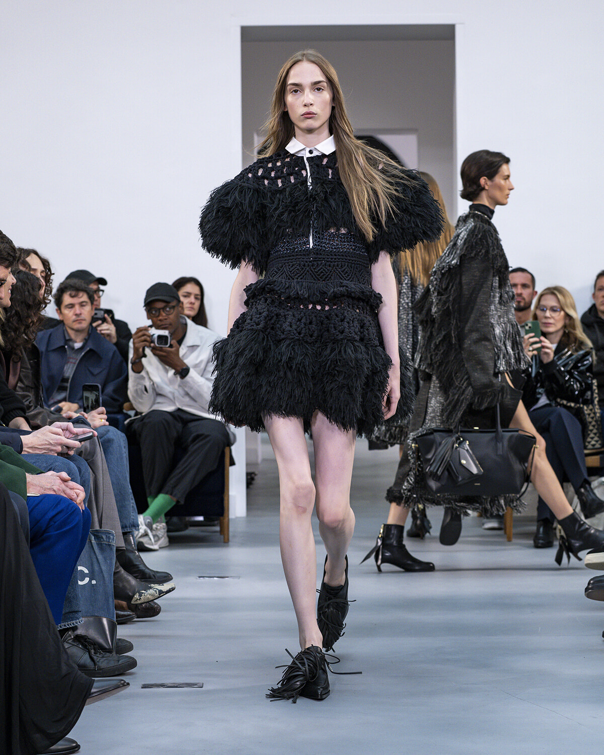 Model walks a runway in a voluminous black feathered dress with a white collar, as spectators watch and photograph the show.