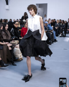Model walking a fashion runway in a white sleeveless blouse and oversized black skirt with fringe shoes; audience seated along the catwalk.