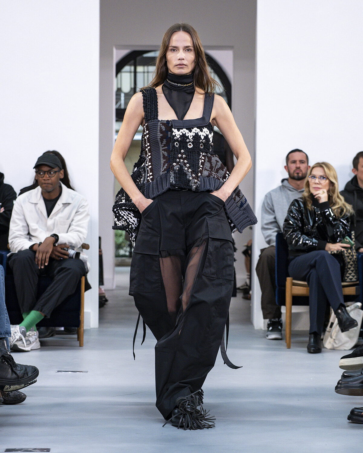 Model walking a fashion runway in a black, patchwork top with varied textures and loose, sheer-panel cargo pants; audience watches from the sides.
