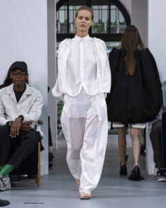 Model walks a runway in an all-white outfit with oversized puff sleeves and wide trousers; audience seated on both sides.