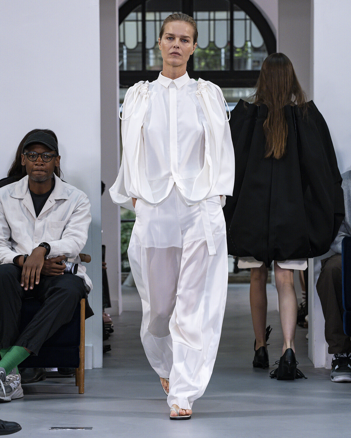 Model walks a runway in an all-white outfit with oversized puff sleeves and wide trousers; audience seated on both sides.