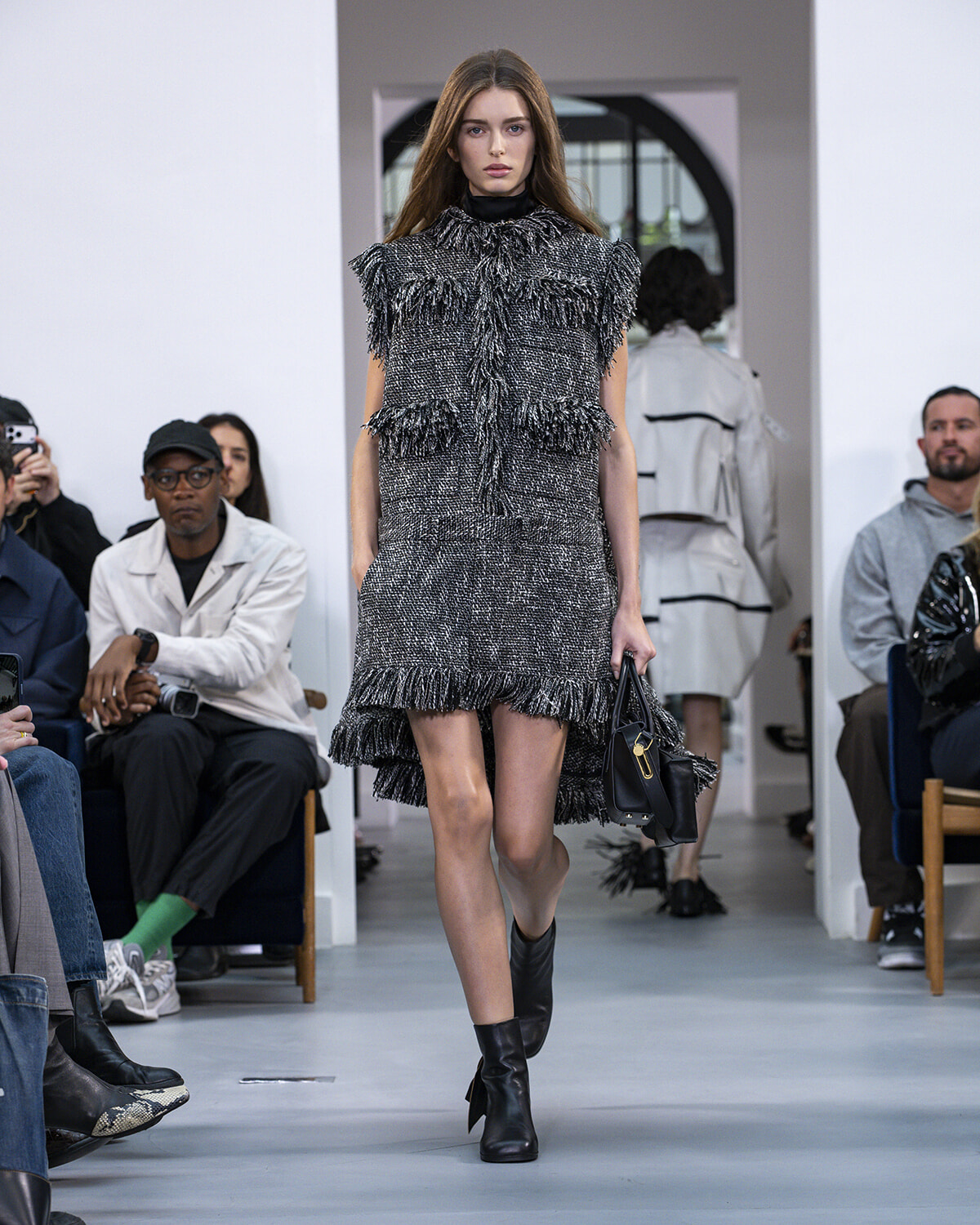 Model walks the runway in a textured black-gray fringe dress with high neck, holding a black handbag and wearing black ankle boots.