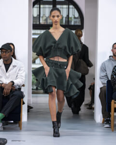 Model walking a runway in a dark green, oversized V-neck top with a matching layered peplum skirt and ankle boots.