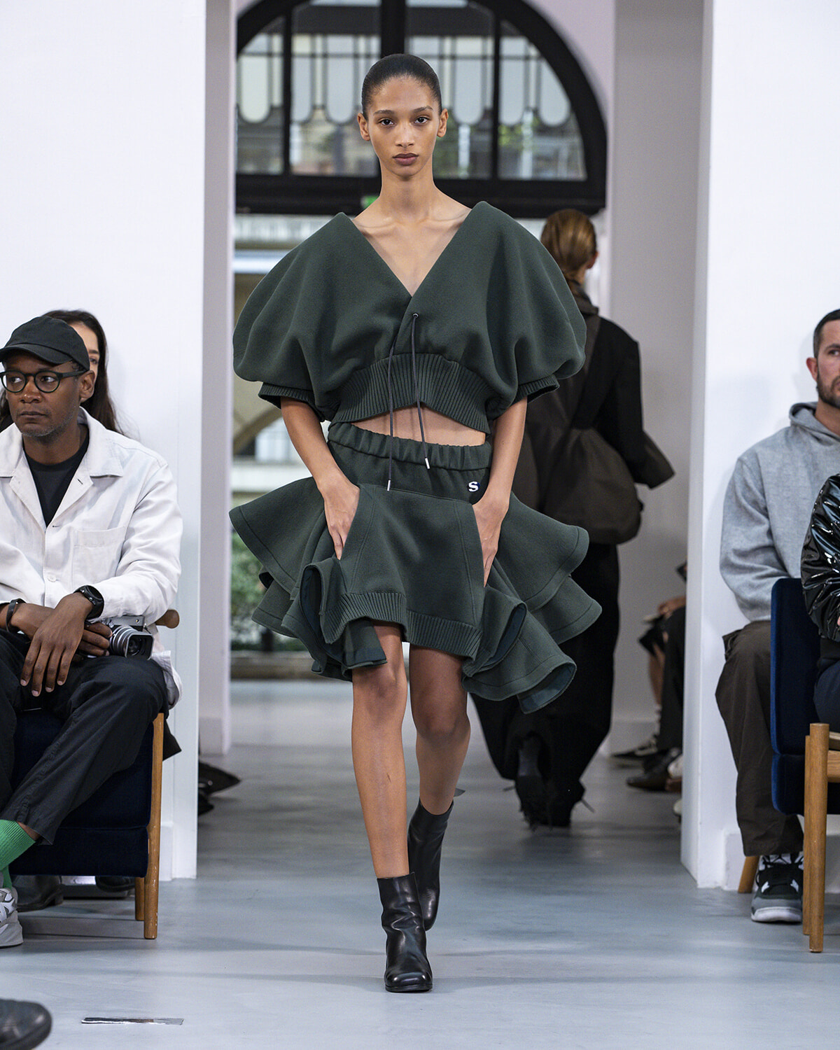Model walking a runway in a dark green, oversized V-neck top with a matching layered peplum skirt and ankle boots.