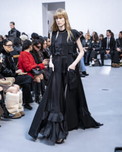Model walks the runway in a long black layered gown with ruffles as a crowd watches from the front row of a fashion show.