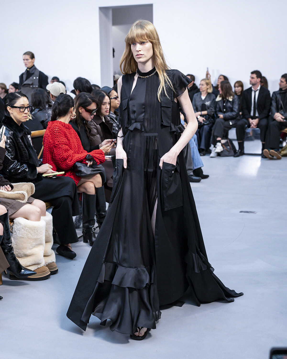 Model walks the runway in a long black layered gown with ruffles as a crowd watches from the front row of a fashion show.